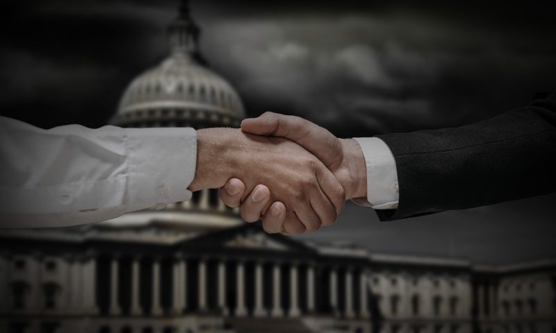 Two hands shaking in front of the United States capitol building with dark clouds.