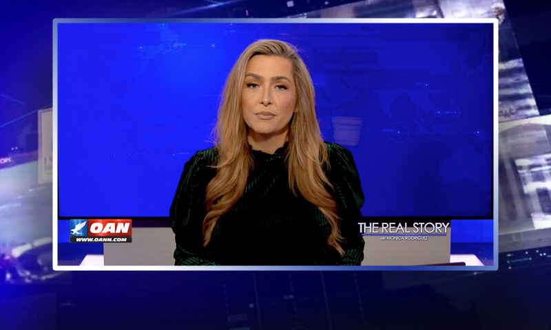 Video still of the host of The Real Story at the desk of their talk show on One America News Network.