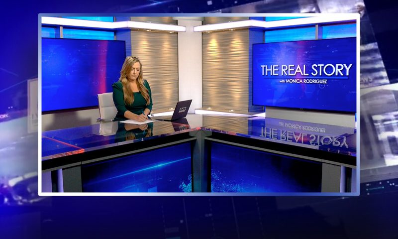 Video still of the host of The Real Story at the desk of their talk show on One America News Network.