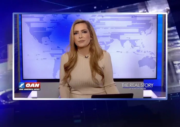 Video still of the host of The Real Story at the desk of their talk show on One America News Network.